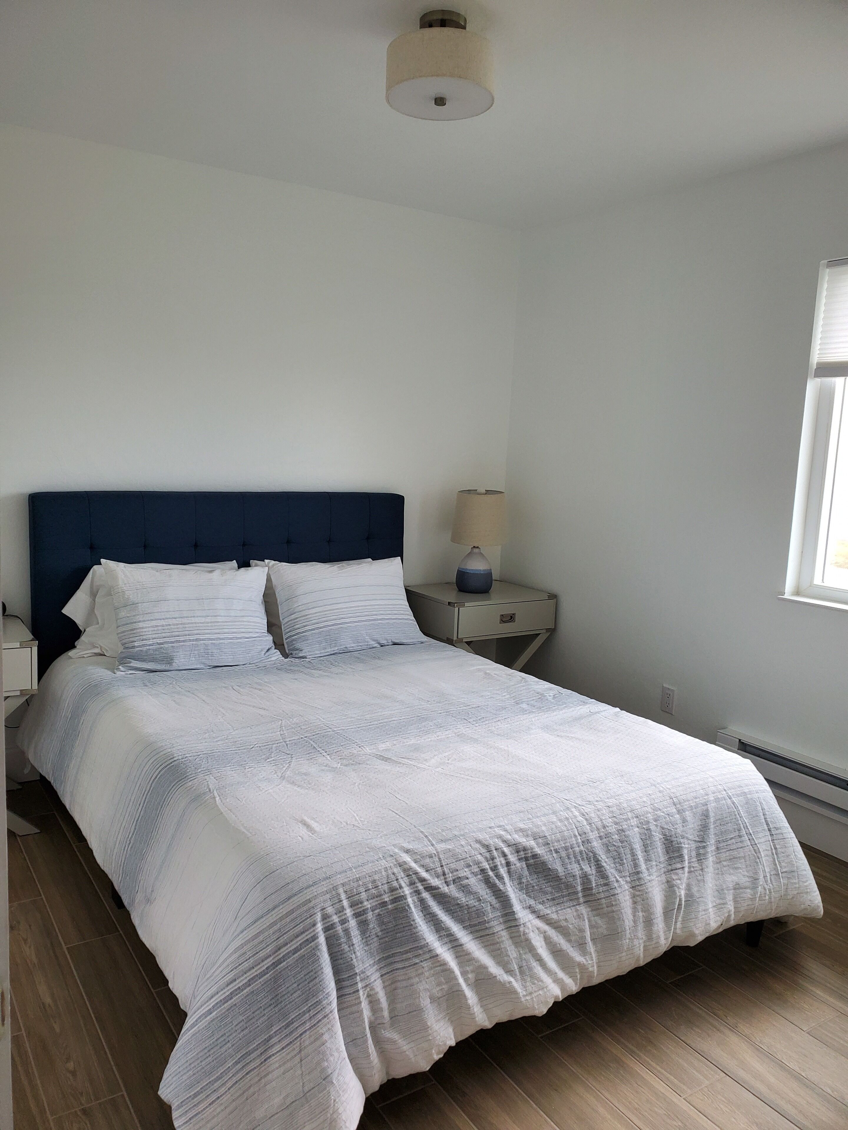 A neatly made bed with a blue headboard, a bedside table with a lamp, and a window with blinds.