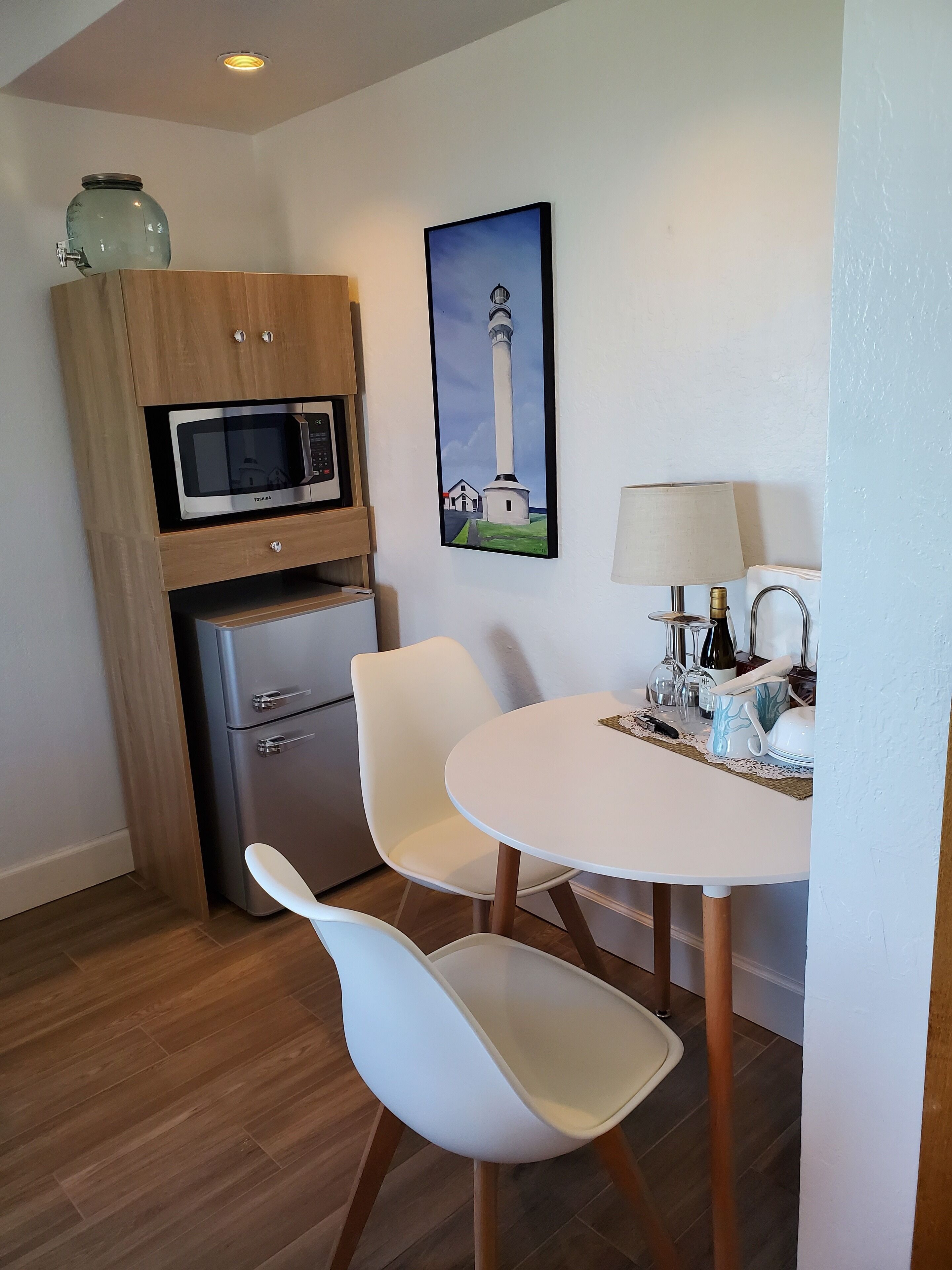 Una cocina compacta con microondas, refrigerador y un mueble pequeño. Hay una mesa de comedor con sillas y un cuadro de un faro.