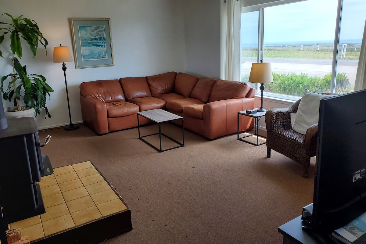 Uma sala de estar com sofá de couro marrom, uma mesa de centro pequena, televisão e uma janela com vista para a praia.