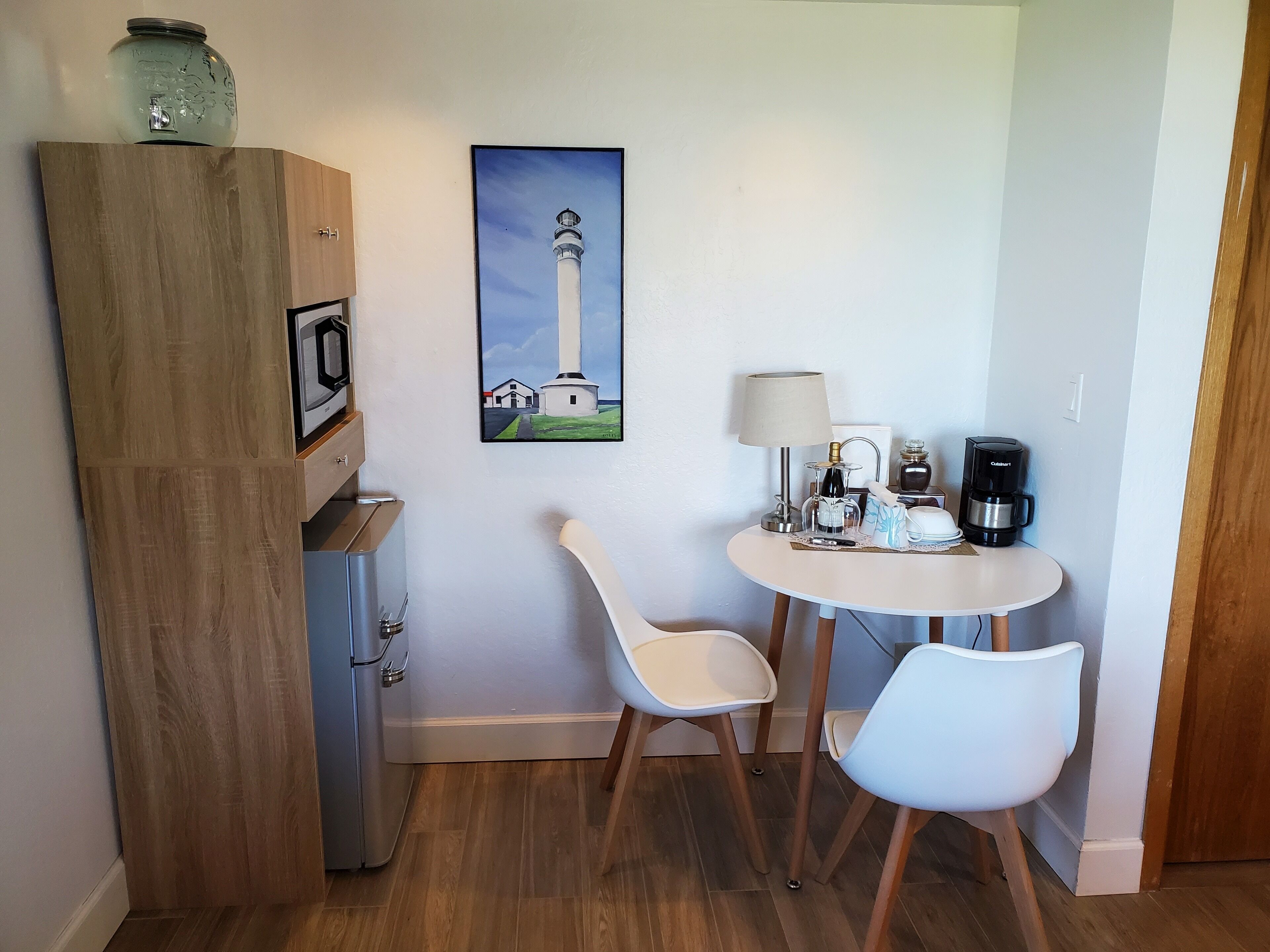 A small room with a wooden cabinet, a refrigerator, a table with two chairs, a coffee maker, and a painting of a lighthouse.