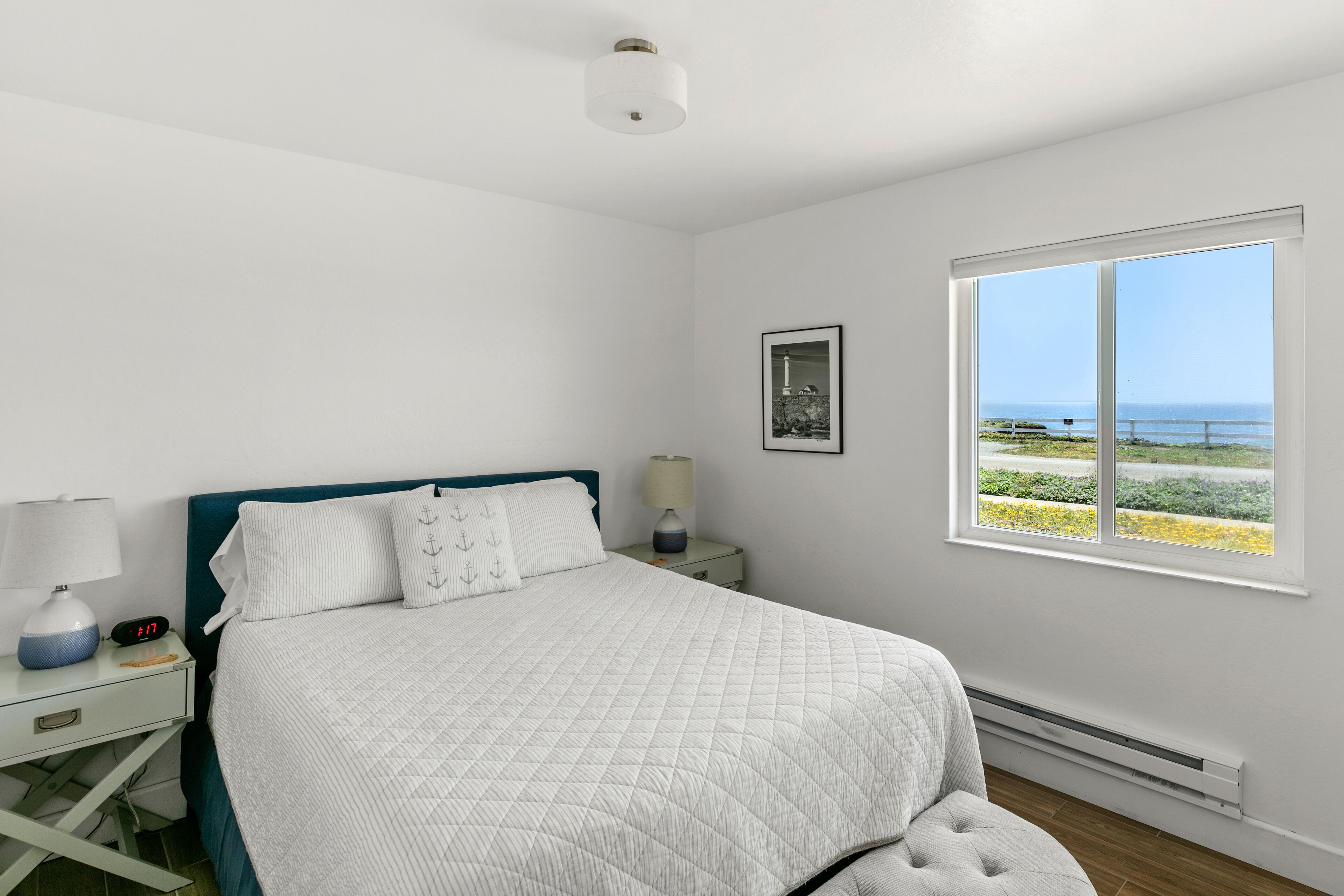 A bedroom with a bed, a nightstand, a lamp, a clock, a picture, and a window with a view of the sea.