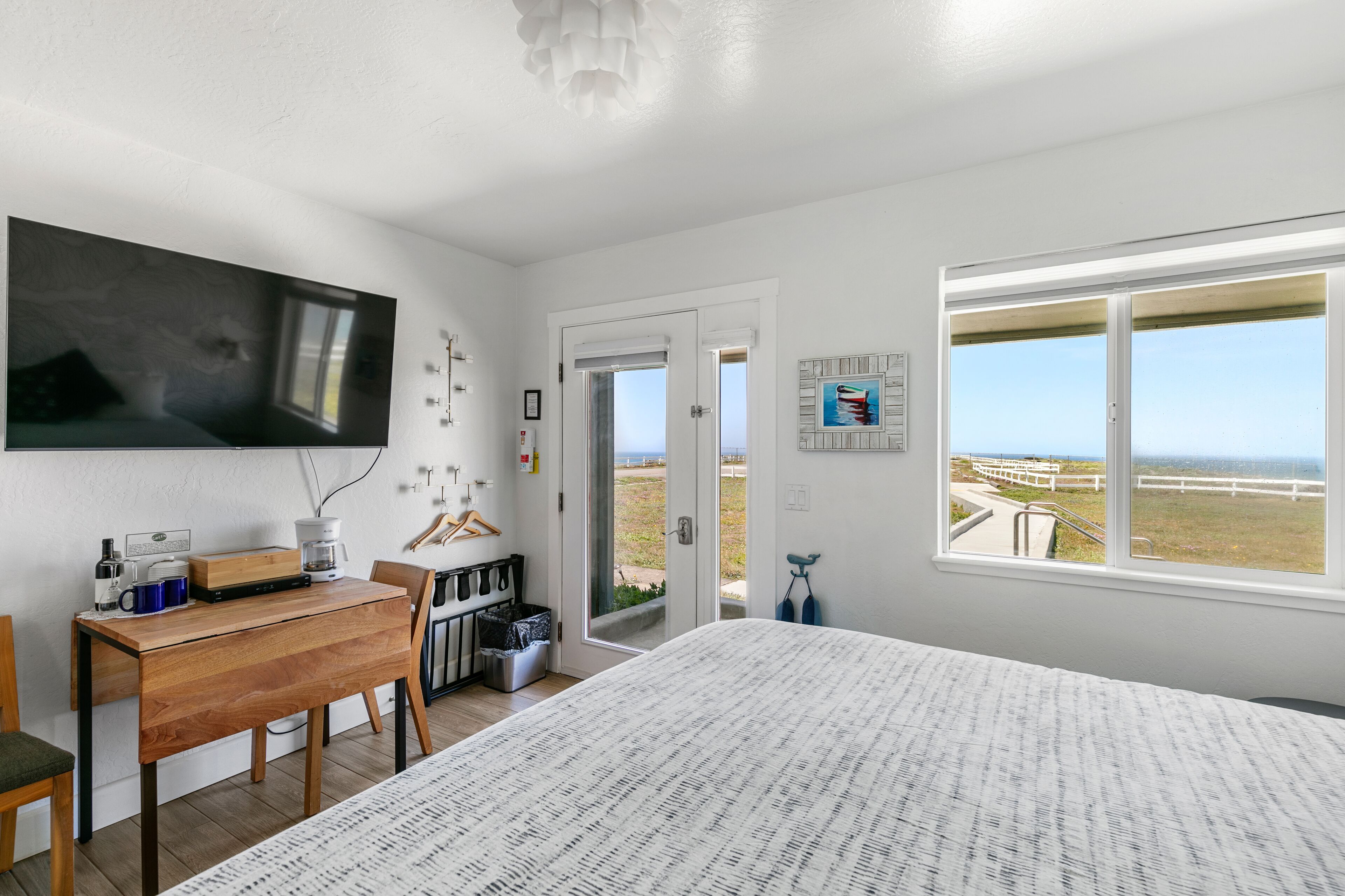 Ett sovrum med en stor säng, ett skrivbord med en dator, en stol och utsikt över stranden genom ett fönster.
