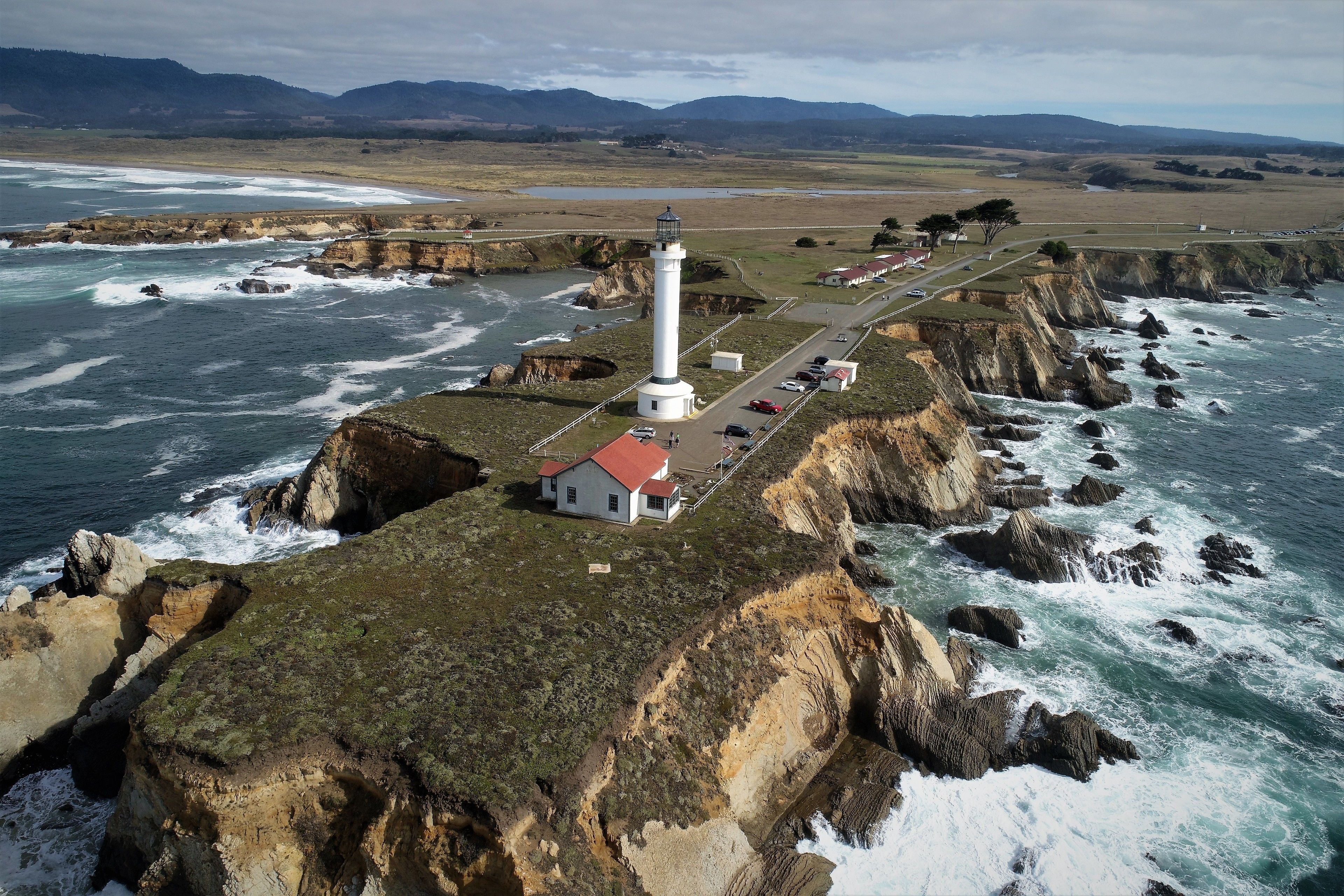 A lighthouse on a cliff with a small building nearby, surrounded by rocky coastline and ocean waves.