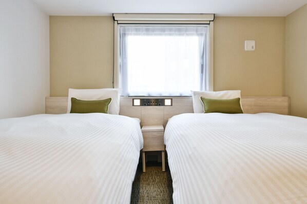 A hotel room with two beds, a window with curtains, a bedside table, and a wall-mounted thermostat.