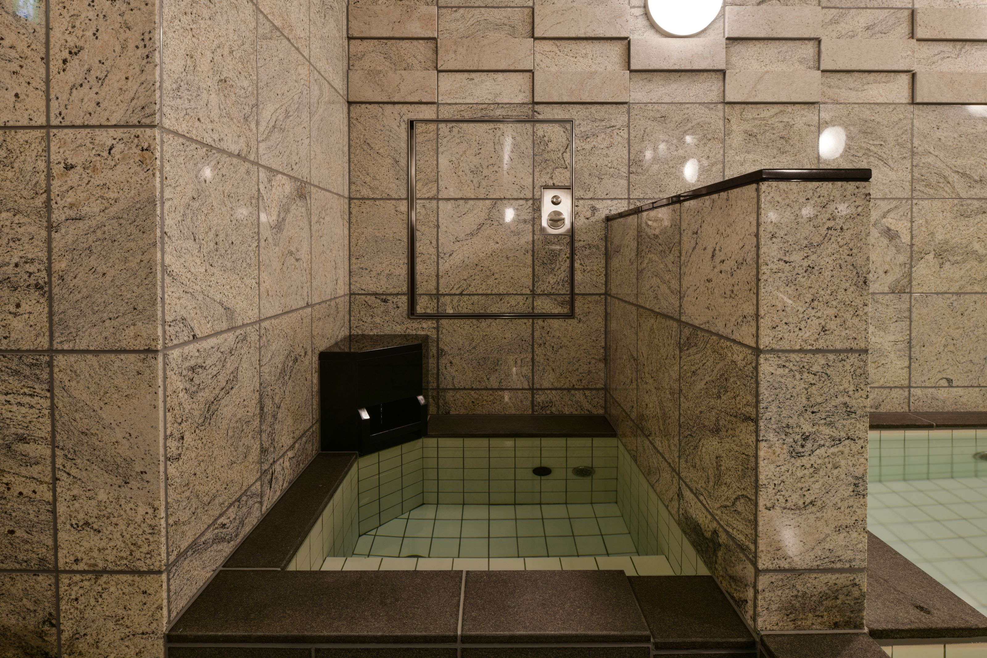 Une petite salle de bain fermée, avec un sol et des murs carrelés, un banc intégré et un luminaire fixé au mur.