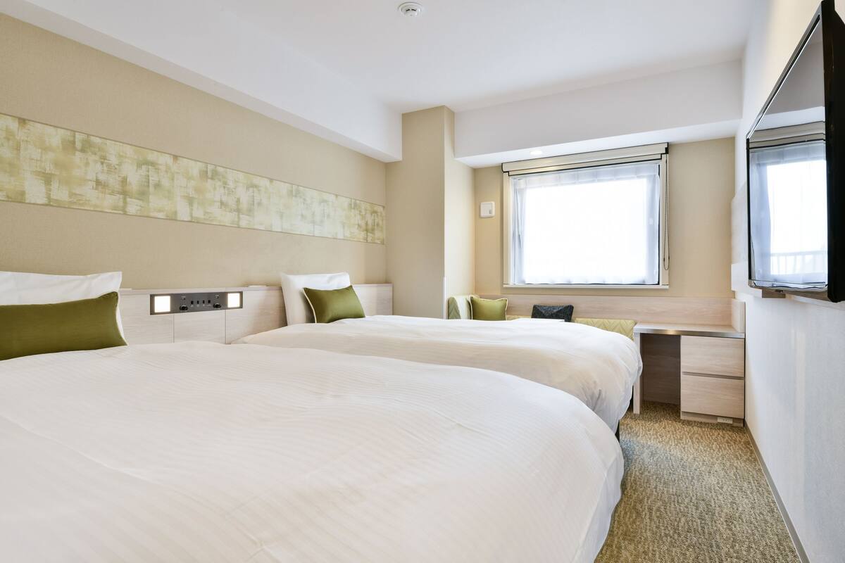 A hotel room with two beds, a desk, a TV, and a window with blinds.