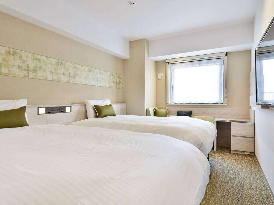 A hotel room with two beds, a desk, a TV, and a window with blinds.