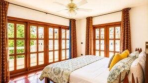 A hotel room with a large bed, wooden flooring, a ceiling fan, and large windows with curtains. - Luxury 6BR Villa / Pool /Walk to Beach (Punta Cana)