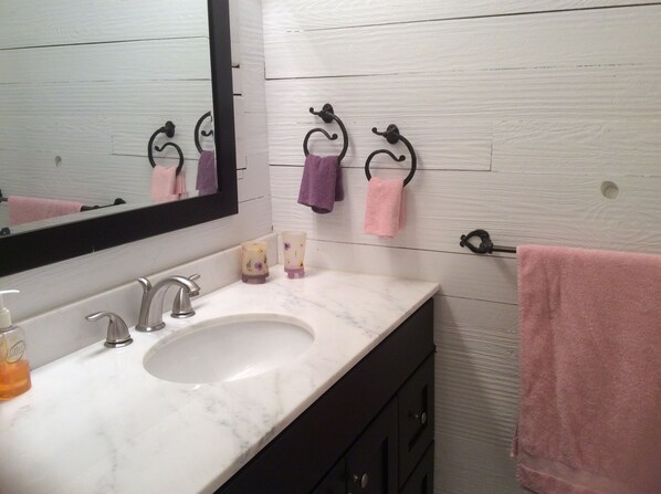 Une salle de bain avec un lavabo en marbre blanc, un grand miroir et un mur en bois. Deux serviettes sont accrochées à des crochets et un flacon de savon est posé sur le comptoir.