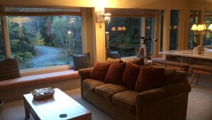 Un salon chaleureux avec un canapé marron, une table basse en bois et une table à manger avec des chaises. De grandes fenêtres offrent une vue sur une zone boisée.