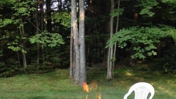 Un foyer en pierre avec un feu allumé, situé dans un espace herbeux, entouré d’arbres et d’une chaise en plastique blanche.