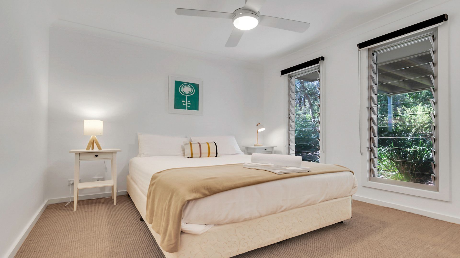 Une chambre à coucher comprenant un lit, des tables de chevet, un ventilateur de plafond et de grandes fenêtres équipées de stores.