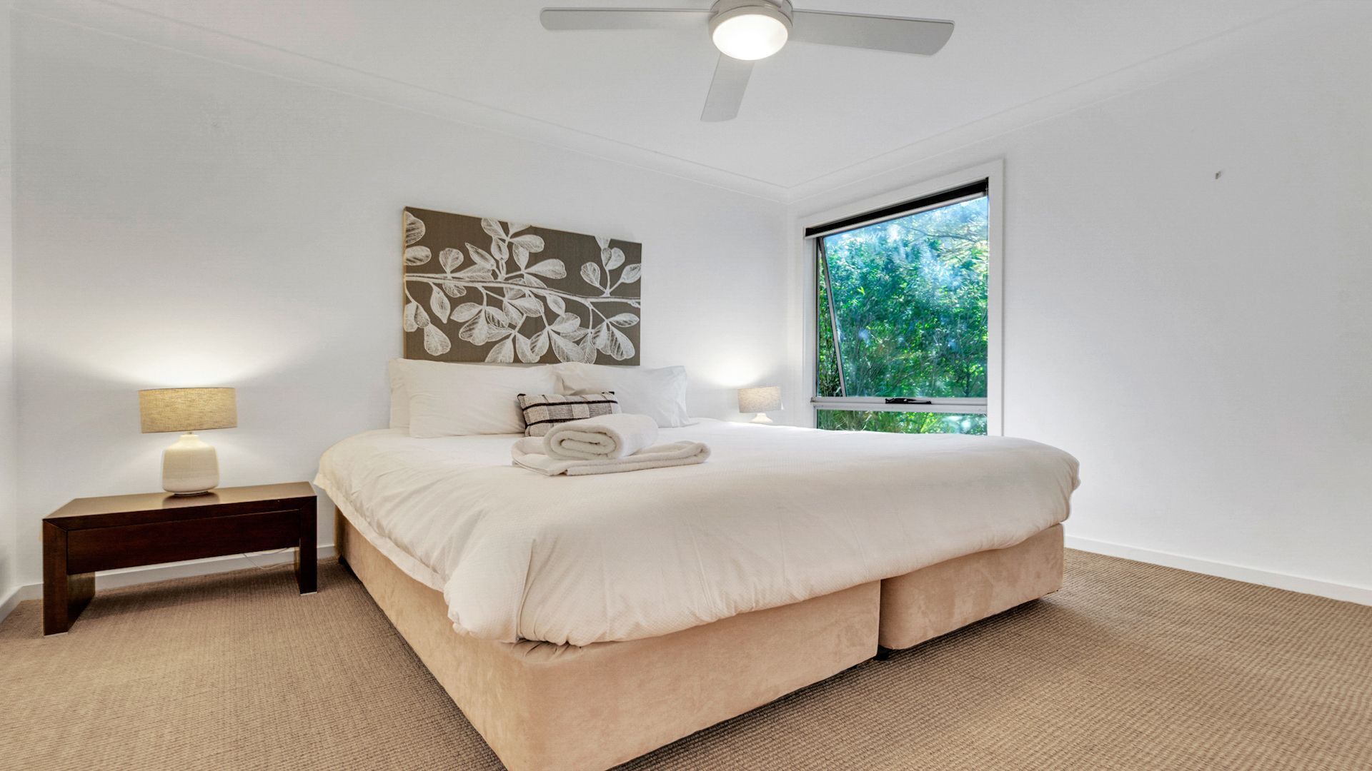 A bedroom with a large bed, two bedside tables, a ceiling fan, and a window with a view of greenery.
