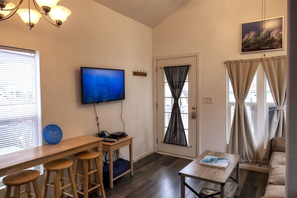 A compact living space with a flat-screen TV mounted on the wall, a wooden bar with stools, a small table with a book, and a door with curtains. - San Juan - Cozy tiny pet friendly home in the heart of the historic Noyo harbor (Fort Bragg)