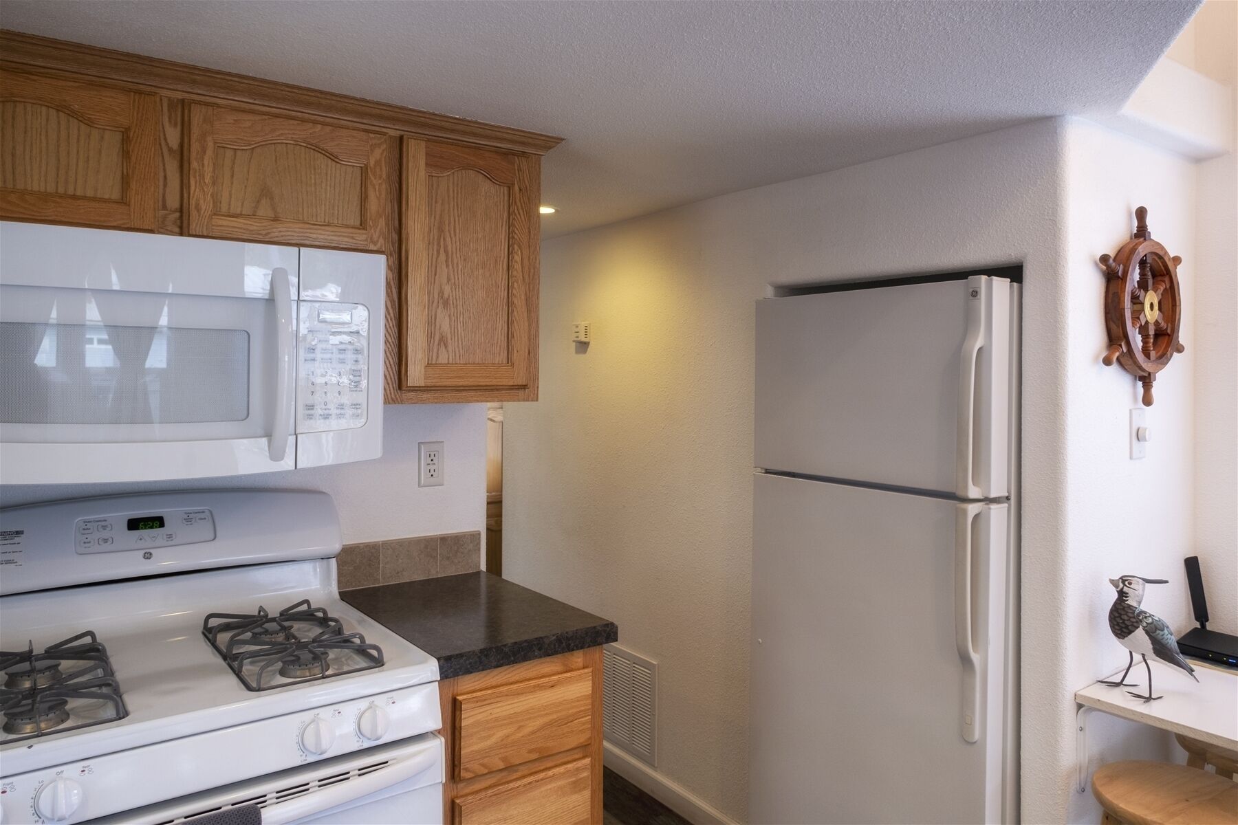 A compact kitchen with a white microwave, stove, and refrigerator. There are wooden cabinets and a small wooden shelf with a bird decoration.
