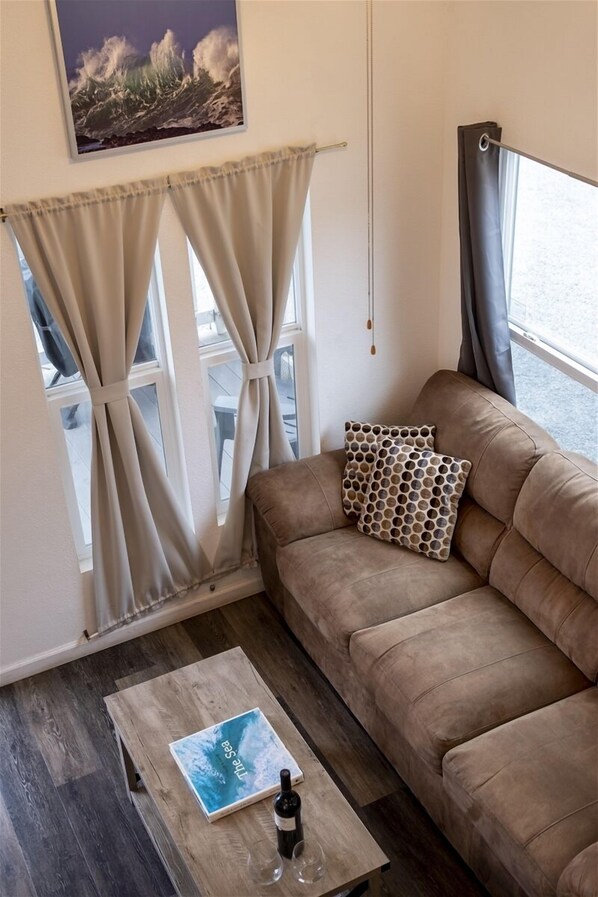 En hyggelig stue med en brun sofa, et træbord med en bog og en flaske, beige gardiner og et indrammet billede på væggen.