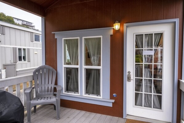 A covered porch with a grey plastic chair, a wooden door with a glass panel, and a window with curtains. - San Juan - Cozy tiny pet friendly home in the heart of the historic Noyo harbor (Fort Bragg)