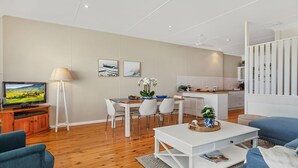Un salon moderne doté d’un téléviseur à écran plat, d’un coin repas avec une table et des chaises, d’une cuisine avec des meubles blancs et d’un escalier avec des rampes blanches.