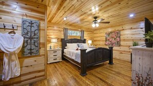 A wooden cabin bedroom with a bed, nightstands, a ceiling fan, and a painting on the wall. - Shear Pleasure|Romantic|Farmhouse Style (Hochatown)