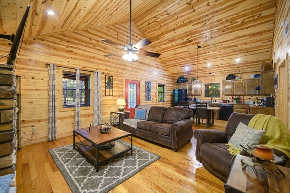 A cozy cabin interior with a wooden ceiling, a dining area, a kitchen, and a living room furnished with a sofa and a coffee table.