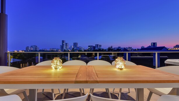 A wooden table with white chairs and two lit candles, set against a city skyline at dusk. - Darwin City Lights Jacuzzi (Stuart Park)
