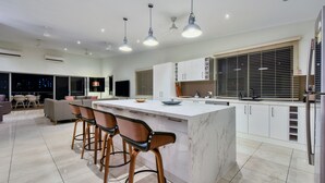 A modern kitchen with a marble island, bar stools, pendant lights, and a large window with blinds. - Darwin City Lights Jacuzzi (Stuart Park)