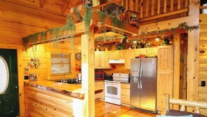 L’intérieur d’un chalet confortable, avec des murs en rondins de bois, une cuisine équipée d’un réfrigérateur, d’une cuisinière et d’un four à micro-ondes, et un chat perché au plafond.