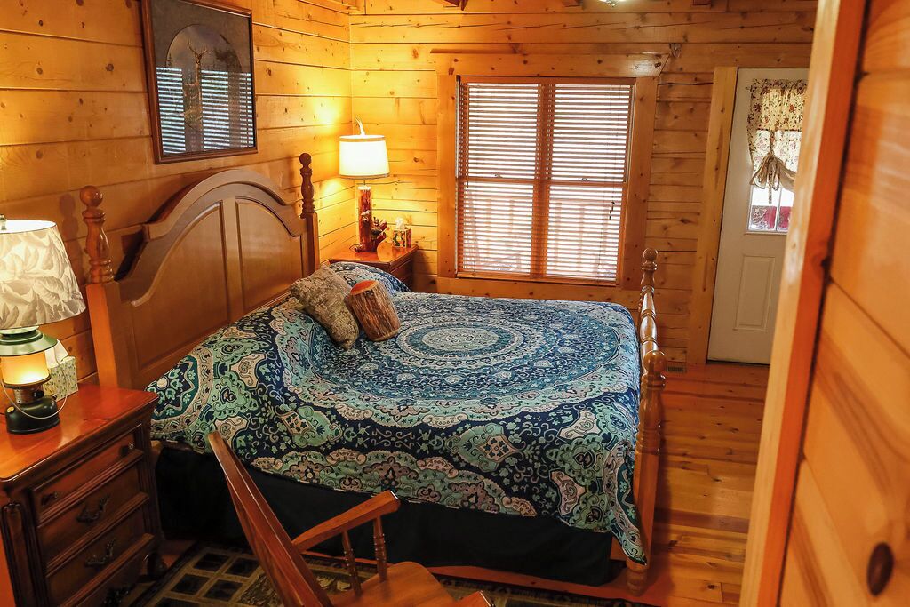 A cozy wooden cabin bedroom with a bed, a nightstand, a lamp, and a window with blinds.