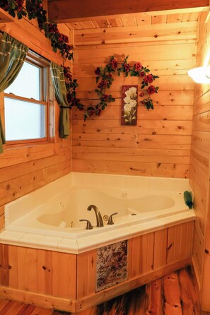 A wooden cabin bathroom with a large bathtub, a window with curtains, and a wall decorated with flowers and a framed picture.
