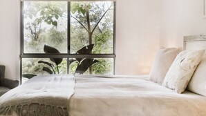 Une chambre avec une grande fenêtre, un lit recouvert de draps blancs et une vue sur les arbres à l’extérieur.