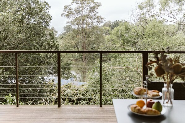 Een balkon met uitzicht op een bebost gebied, een tafel met een bord eten en een vaas met gedroogde bloemen.