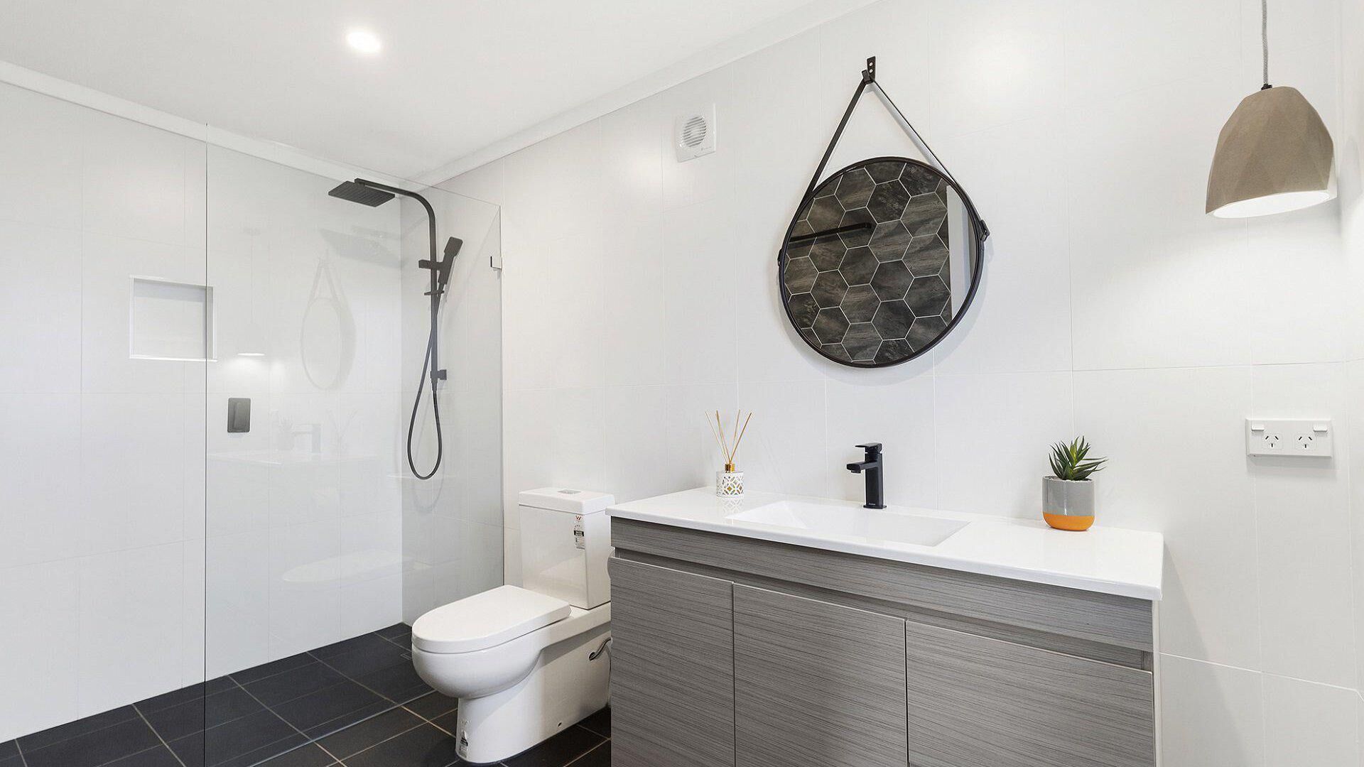 Un baño moderno con ducha, inodoro, lavabo y un espejo redondo con un revestimiento de azulejos hexagonales.