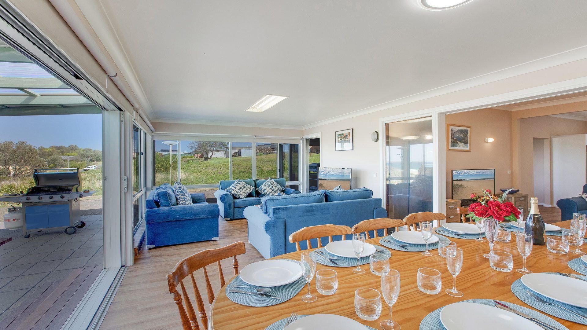 A spacious living area with a dining table set for six, blue sofas, and a view of the outdoors.