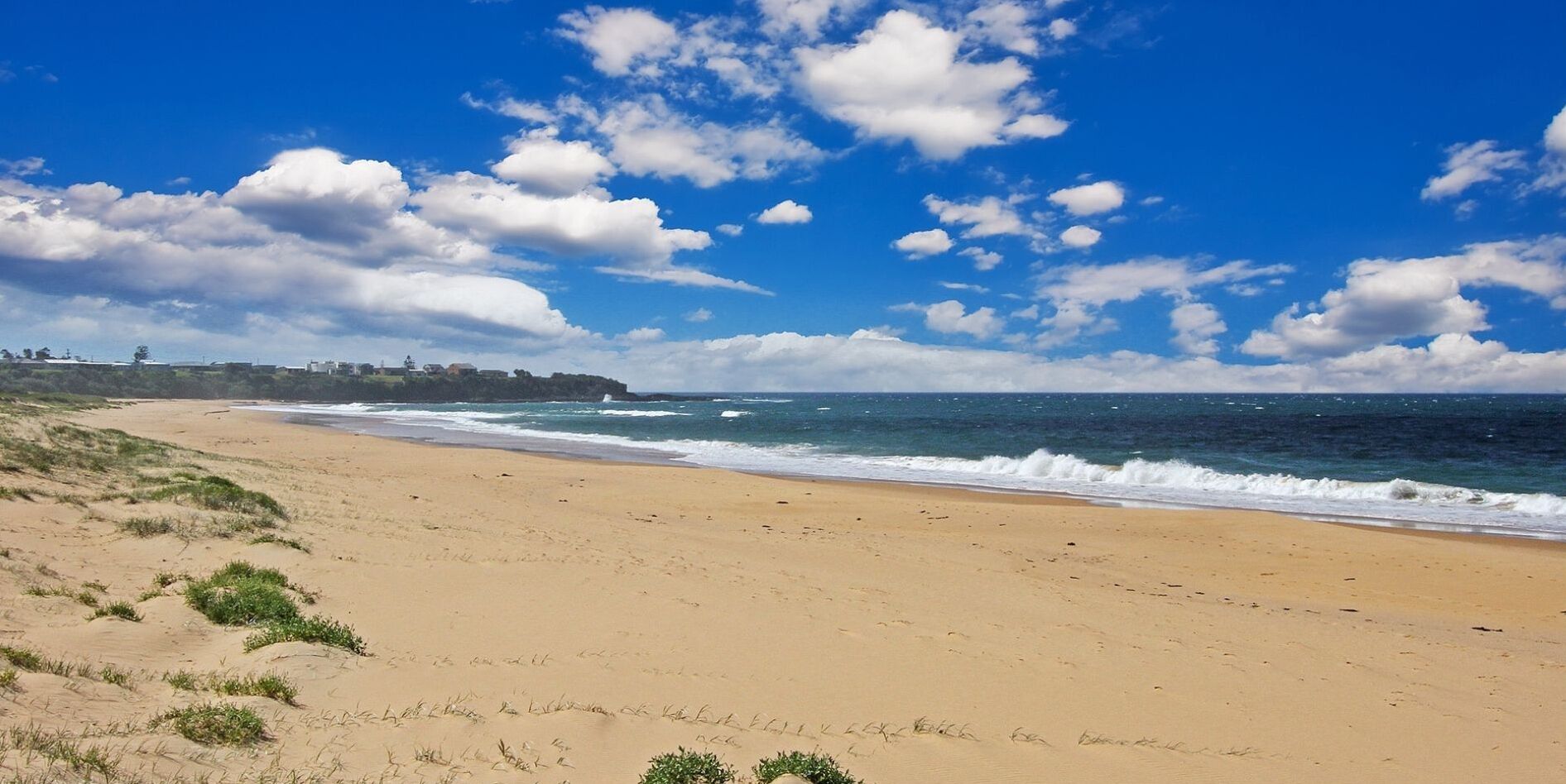 Une vaste plage de sable fin, avec des vagues douces, un ciel bleu clair et quelques nuages épars.