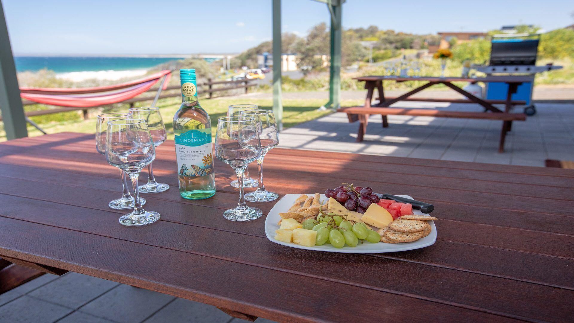A wooden table with a plate of cheese, grapes, and crackers, a bottle of wine, and three wine glasses.