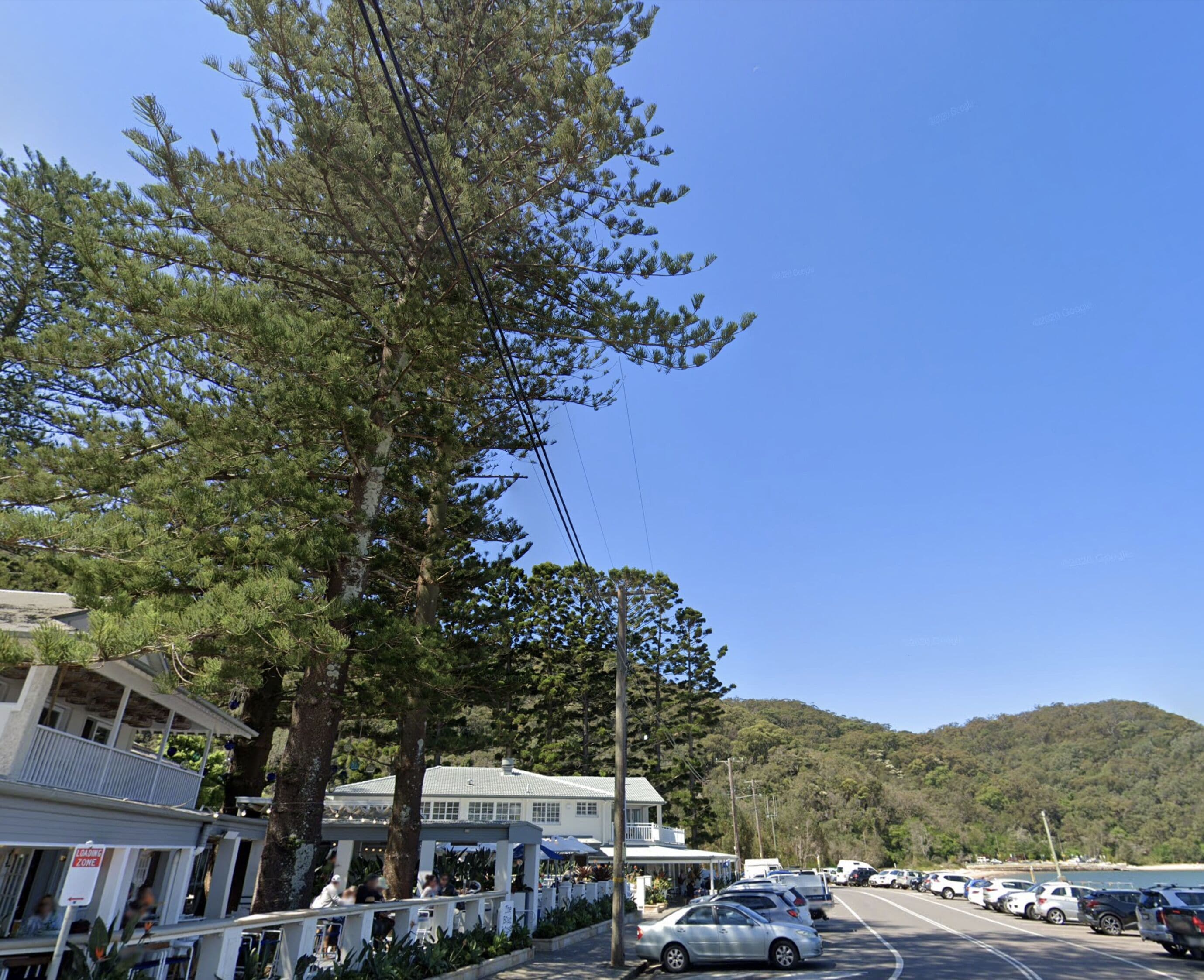 A coastal road with parked cars, a white building with a balcony, and a forested hill in the background.