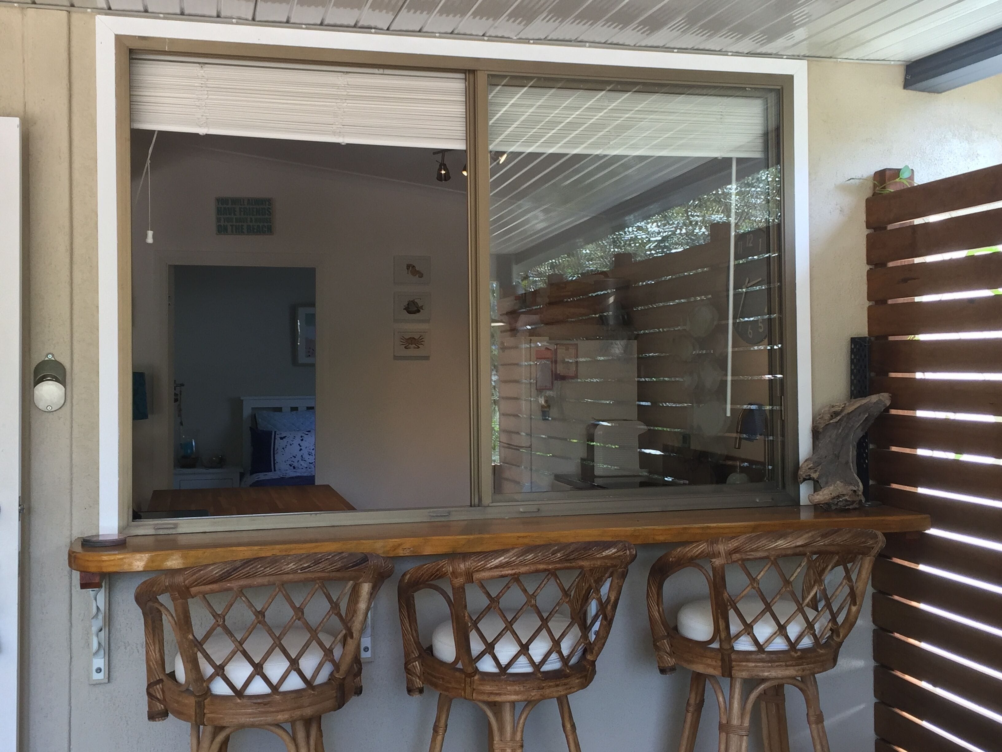 A wooden bar with three wicker chairs, a sliding glass door leading to a room, and a wooden slatted wall on the right.