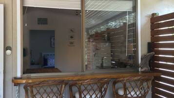 A wooden bar with three wicker chairs, a sliding glass door leading to a room, and a wooden slatted wall on the right.
