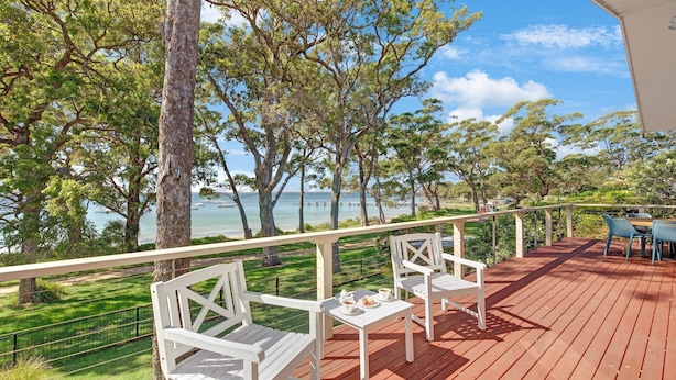 A wooden deck with white chairs and a table, overlooking a body of water with boats.