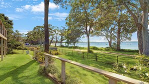 A grassy area with a wooden fence, trees, and a view of the sea with boats.