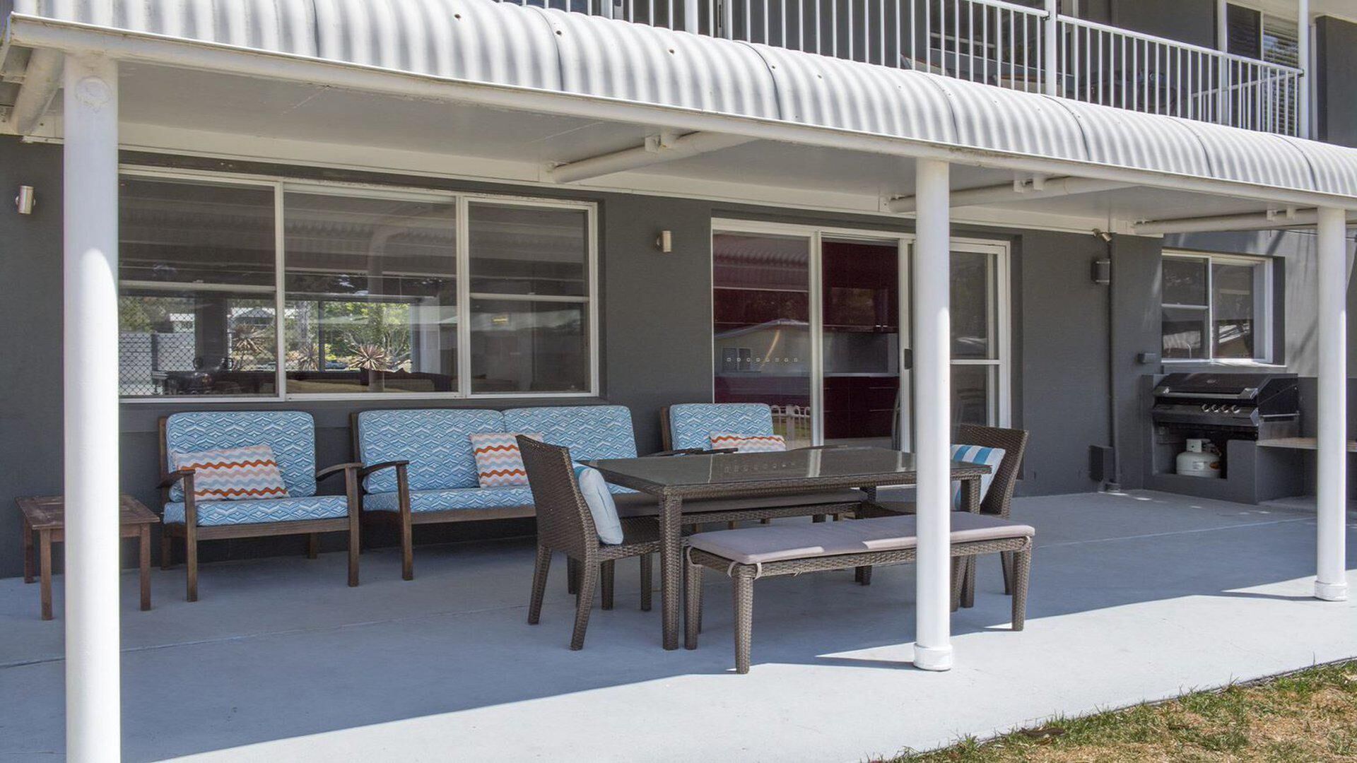 Une terrasse couverte avec une table à manger et des chaises, un barbecue et un coin salon agrémenté de coussins à motifs.