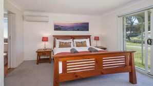 A bedroom with a wooden bed, two bedside tables with lamps, a painting on the wall, and a view through a glass door. - Kendalls Beach Escape (Kiama)