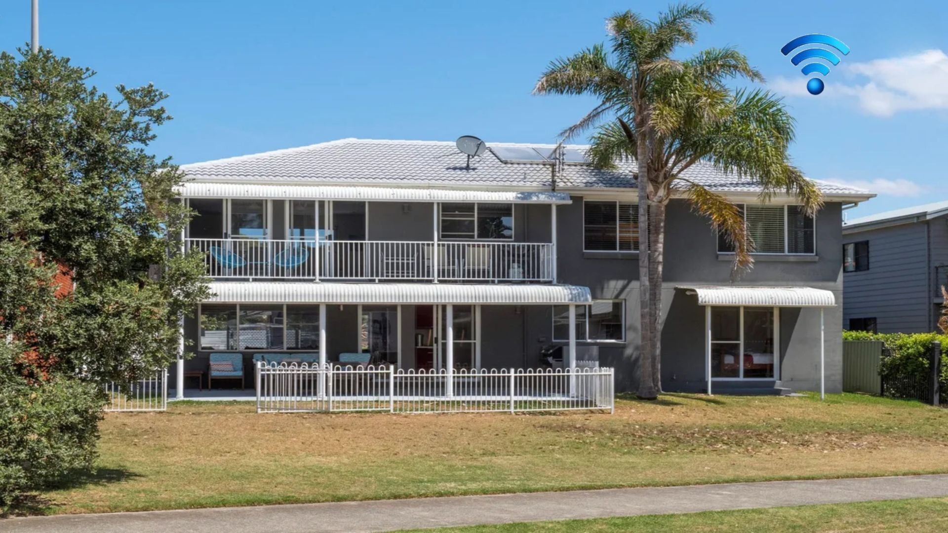 A two-story residential building with a balcony, a satellite dish, and a palm tree in the yard.
