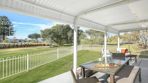 A covered outdoor area with a table and chairs, a grassy lawn, and a white fence. - Kendalls Beach Escape (Kiama)