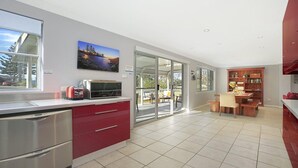 A modern kitchen with a sliding glass door leading to a patio, a dining area with a wooden table, and a red cabinet. - Kendalls Beach Escape (Kiama)