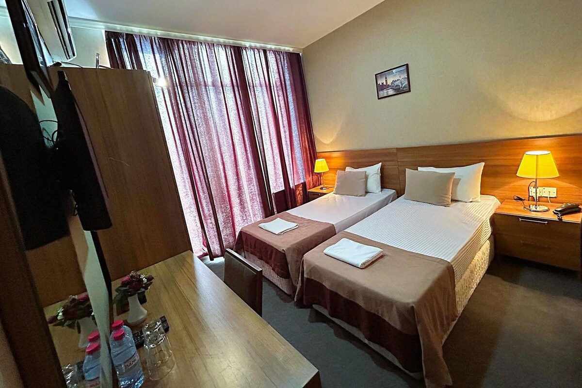 A hotel room with two beds, a desk, and a chair. There are two lamps on the nightstands, a picture on the wall, and a window with curtains.