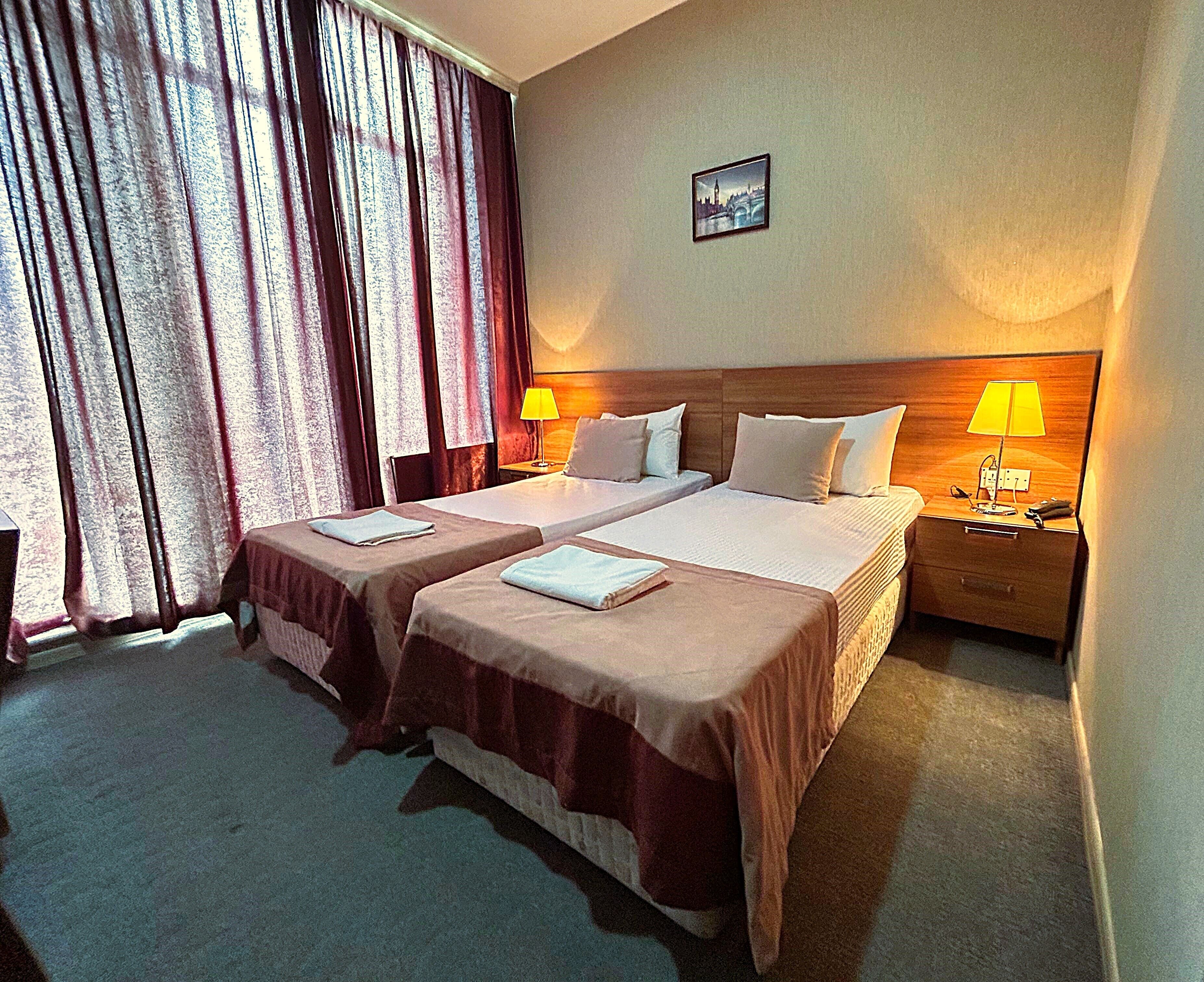 A hotel room with two beds, a nightstand with a lamp, a picture on the wall, and a window with curtains.