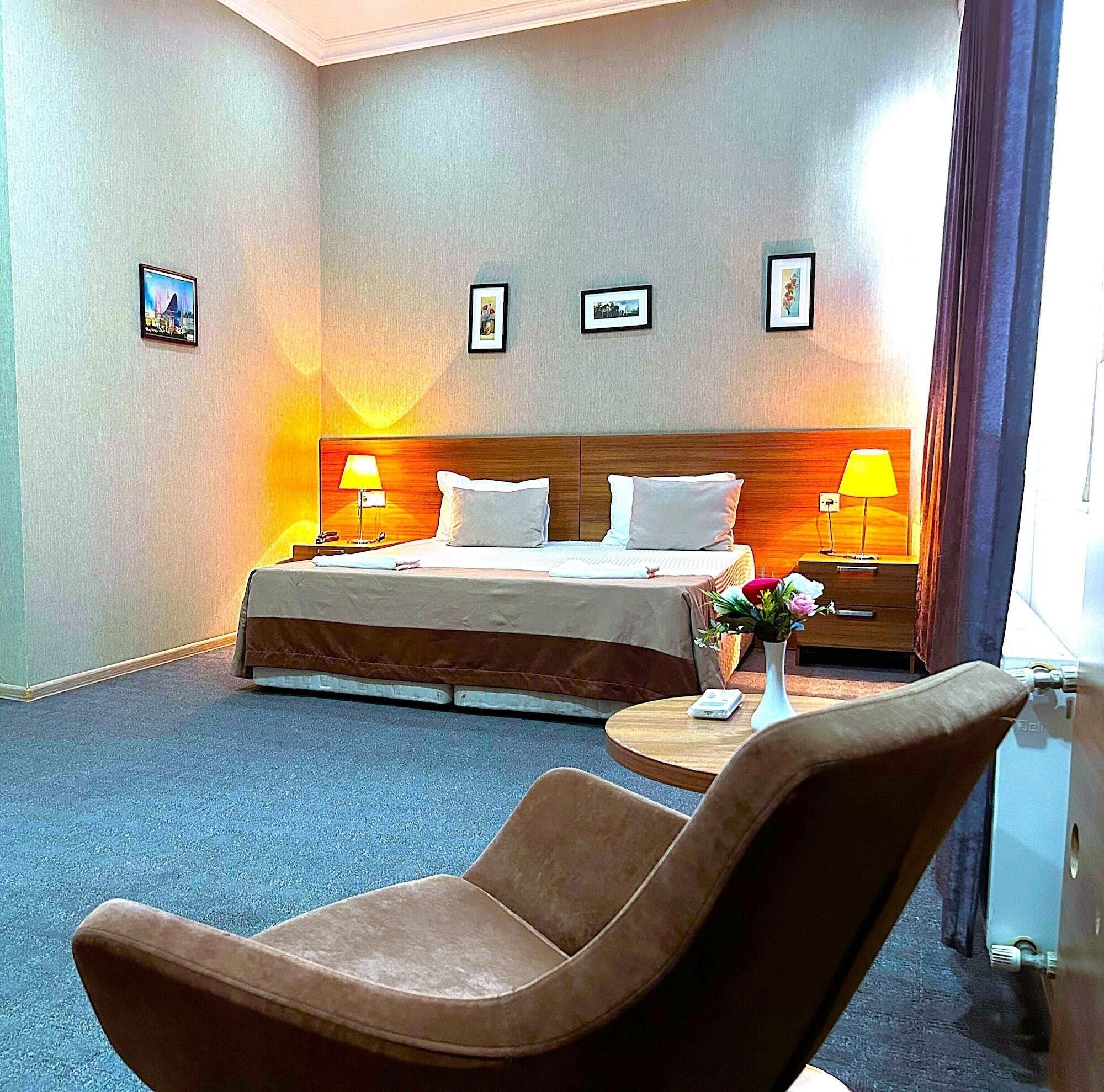 A hotel room with a bed, two bedside lamps, a chair, a small table with a vase, and three framed pictures on the wall.