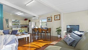 A spacious living area with a sofa, a dining table, and a kitchen in the background. - Between 2 Beaches – pet friendly cottage (Culburra Beach)