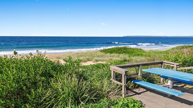 A wooden bench and picnic table set on a concrete path with a view of the ocean and beach vegetation. - Between 2 Beaches – pet friendly cottage (Culburra Beach)